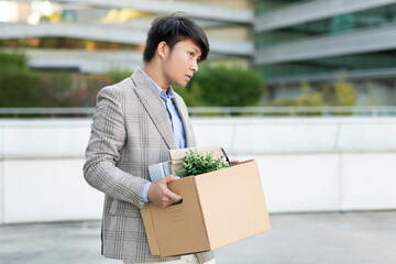 A young Asian man walks away from a modern office building, carrying a cardboard box filled with personal items. He appears dejected and downtrodden, perhaps having just been laid off or resigned