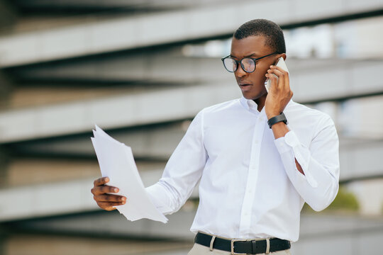 African American businessman in a white shirt and glasses is standing outside, holding papers in his left hand and talking on his phone with his right hand. He is looking down at the documents - Powered by Adobe