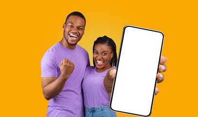 African American young couple, dressed in purple shirts, stand in front of a solid yellow background. The woman, holding a smartphone with a blank screen, points it towards the camera