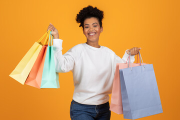 A young woman with short, curly hair is smiling and holding colorful shopping bags in each hand. She is wearing a white long-sleeved shirt and blue jeans and standing in front of yellow background.