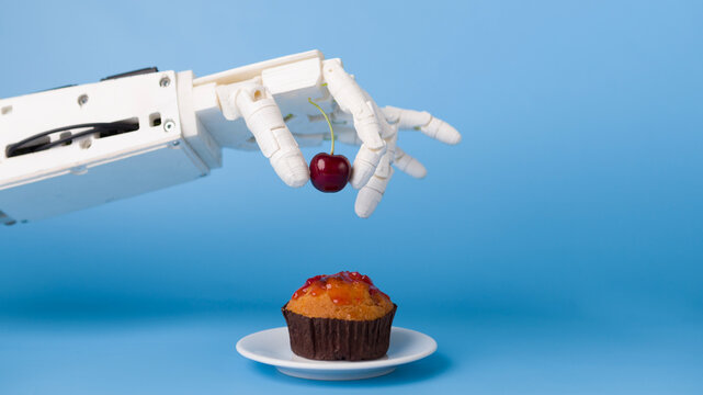 Kitchen robotization. Robot hand putting fresh cherry on top of the cupcake, blue background