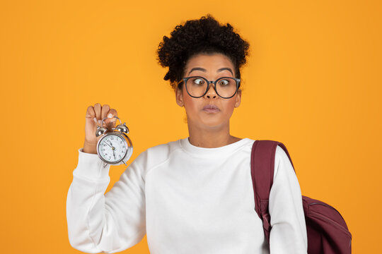 A young woman with glasses and a backpack stares in surprise as she holds a silver alarm clock. The clock is in her right hand, held out in front of her, posing against a solid yellow background.