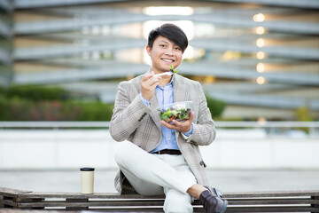 Asian man in a blazer sits on a bench outside an office building. He eats a salad with a fork and smiles.