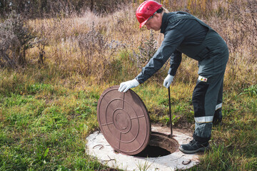 A worker in a helmet lifts a manhole cover to service a well. Plumbing and sewerage