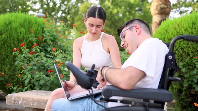 A young girl cares for an adult man with cerebral palsy in a park at sunset.The young woman helps the disabled man work with a laptop.Concept of people with dependence due to cerebral palsy.