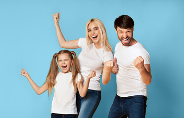 A family of three, a young girl, a woman, and a man, stand together in front of a solid blue background, all raising their fists in the air and smiling with excitement.