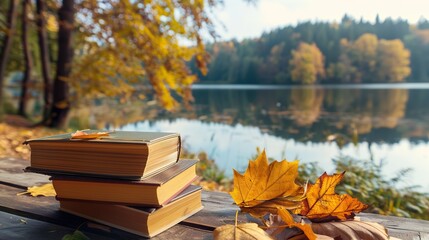 A stack of books on a wooden surface with autumn leaves scattered around, overlooking a calm lake surrounded by trees with autumn foliage.