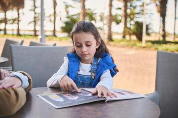 Young girl reading a menu at an outdoor café with sunny weather and palm trees in the background