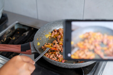 Handphone recording to woman influencer host cooking spaghetti with meat topped tomato sauce surrounded ingredients recipe, presenting special dish healthy food at modern studio.