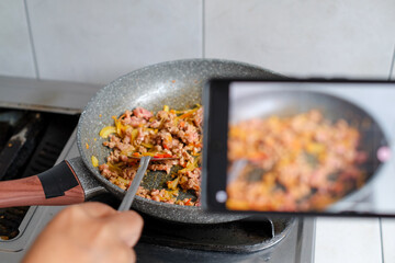 Handphone recording to woman influencer host cooking spaghetti with meat topped tomato sauce surrounded ingredients recipe, presenting special dish healthy food at modern studio.