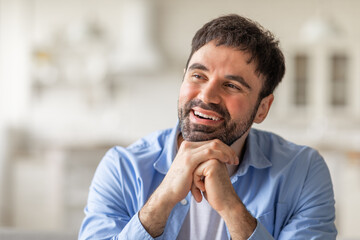 A smiling man with a beard rests his chin on his hands and looks thoughtfully toward copy space, possibly lost in thought or daydreaming