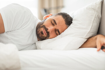 A man with a beard sleeps peacefully on his side, his head resting on a white pillow and his arm...