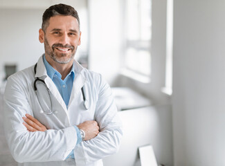 Portrait of friendly middle aged european male doctor in workwear with stethoscope on neck posing with folded arms in clinic interior, looking and smiling at camera, free space