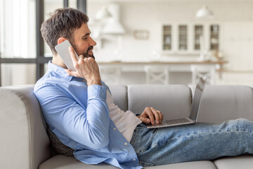 A man wearing a blue button-down shirt and jeans sits on a gray couch in a living room. He is talking on a white smartphone and working on a laptop computer.