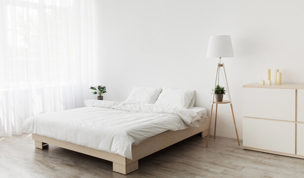 Simple modern design, ad, offer. Double bed with white pillows and soft blanket, lamp, furniture on wooden floor. White empty wall, big window with curtains in bedroom interior