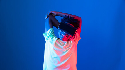 Young African American sport player wearing VR stretching arm at top body posing on blue pink neon light wall connecting digital futuristic technology virtual reality at metaverse world. Contrivance.