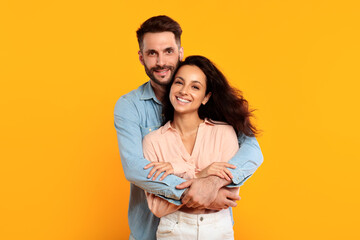 A man with a beard is embracing a woman with long dark hair. They are both smiling and looking directly at the camera. They are standing in front of a solid yellow background.