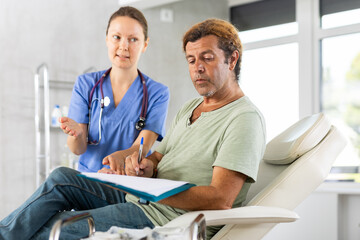 Focused male patient signing informed consent form while lying on couch during medical consultation with female doctor. Compliance with legal requirements when receiving personalized medical care