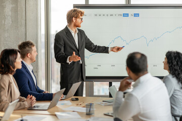 A businessman, wearing a suit, stands in front of a large screen displaying a financial chart, gesturing towards the data. He is presenting to a group of colleagues, who are seated at a table