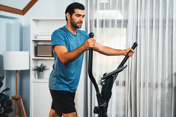 Athletic and sporty man running on elliptical running machine during home body workout exercise session for fit physique and healthy sport lifestyle at home. Gaiety home exercise workout training.