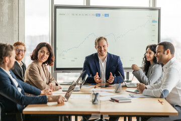 A team of five business professionals sit around a large wooden table in a modern office. The man at the head of the table is presenting a large graph on the screen behind him
