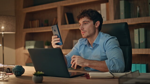Freelancer looking cellphone drinking coffee at work desk late evening close up.