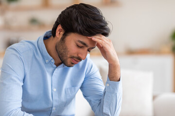 Arab man with dark hair is sitting in a chair indoors. He has a stressed expression on his face. He is holding his hand to his forehead, as if he is experiencing a headache or feeling overwhelmed.