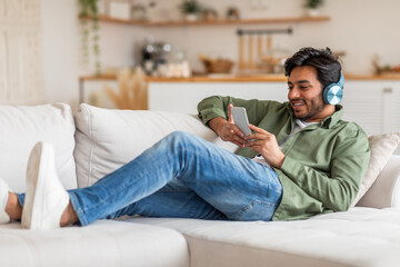 Obraz premium Arab man is lying on a white couch, wearing jeans and a green shirt. He has headphones on and is looking at his smartphone with a smile. He appears to be relaxing and enjoying his time.