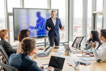 A group of professionals are gathered around a table in a modern office, listening to a businessman who is standing in front of a large screen displaying a graph showing upward growth.