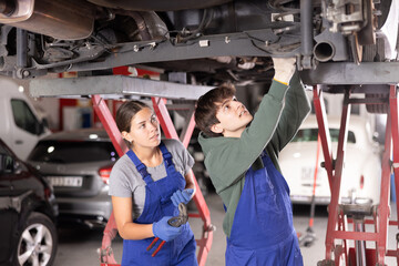 Young guy and young woman mechanics in uniform repairing underbody of car in car service station