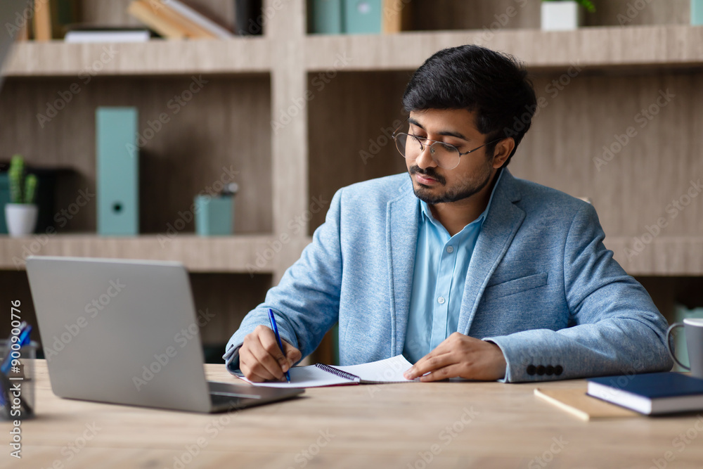 Aufkleber indian man in a blue blazer and glasses sits at a wooden desk, writing in a notebook with a blue pen