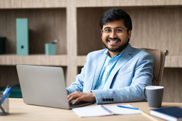 A young Indian man in a blue suit sits at a desk in an office, smiling as he types on a laptop computer. He has a cup of coffee beside him, and there are papers and a notepad on the desk.