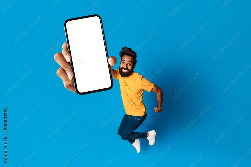 Poster indian man with a beard is jumping in the air, smiling, and holding a smartphone in front of him wit
