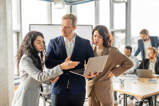Three business professionals stand together in a modern office, engaged in a collaborative discussion while looking at a laptop. The man in the center appears to be leading the conversation - Powered by Adobe