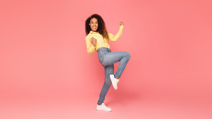 A young black woman in a yellow shirt and blue jeans is dancing in a studio with a pink background. She has her right leg bent at the knee, her left leg straight, and is raising her arms in the air