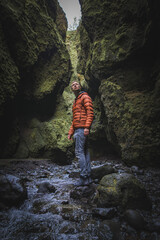 Obraz premium Individual in orange jacket and gray pants stands in a narrow, moss-covered canyon with towering rock walls and a small stream, showcasing Iceland's natural beauty.
