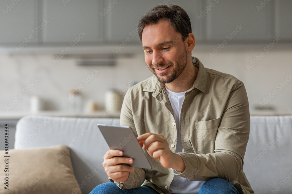 Poster a man sits on a couch in a living room, smiling as he uses a tablet computer. he is wearing a beige 