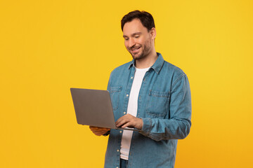 A man wearing a blue denim shirt stands against a yellow background, smiling as he uses a laptop computer.