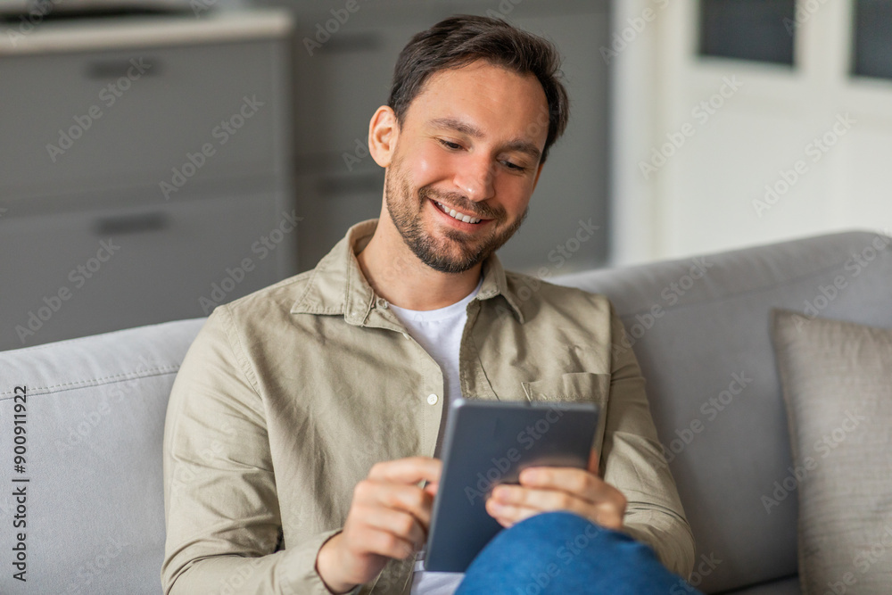 Poster a man sits on a gray couch in a living room, smiling while using a tablet. he is wearing a beige but