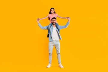 Fototapeta premium A father and his daughter are laughing together as she sits on his shoulders and holds his hands while they stand in front of a vibrant yellow background. They appear to be enjoying a playful moment.