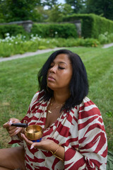 Woman sitting in the grass in a park holding a meditation bell singing bowl. 