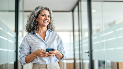 Happy middle aged business woman holding mobile cell phone using cellphone in office. Smiling mature old professional lady business investor owner entrepreneur using smartphone standing looking away.