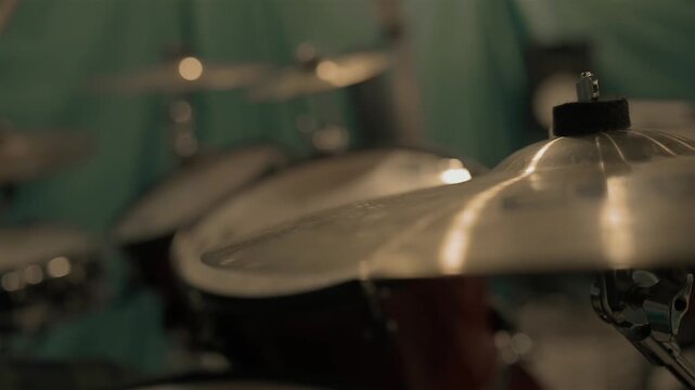 A drummer plays their kit in slow-motion, with a shimmering cymbal in focus in the foreground. The background remains blurred as the performer strikes other drums, creating a dynamic visual rhythm. 