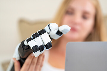 A young woman is using a robotic prosthetic arm hand attached to her body to work on a desk. she is...