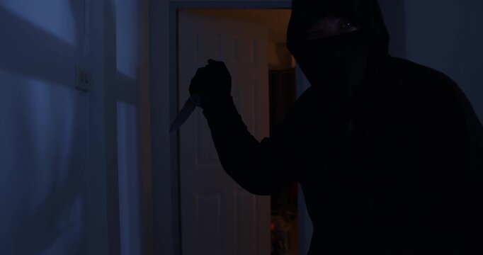 A thief in a black cloak and gloves sneaks into a house at midnight, holding a knife and flashlight, with the intent to harm the occupants.
