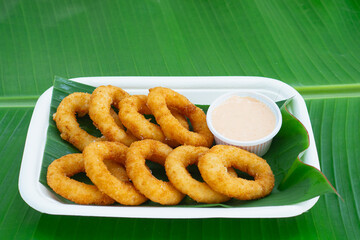 Golden onion rings served with sauce in biodegradable tray on green leaf.