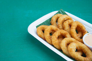 Tasty onion rings served on biodegradable tray with green leaf and sauce.