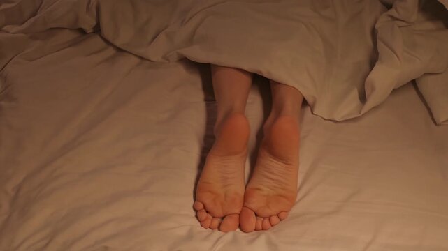 female legs joyfully movie and dance with toes while lying in bed on a sheet under a blanket. Happy woman girl with bare legs foot is relaxing and rest on a bed in a hotel bedroom in the evening