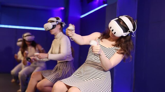 Group of excited young girls sitting with handheld controllers in hands engaged in virtual world experiencing through VR glasses covering face. Concept of immersion and interactivity in game