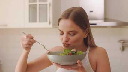 The woman is greedily eating salad in a modern kitchen. She is eating from a bowl filled with greens and cherry tomatoes, holding a fork in one hand and the bowl in the other.